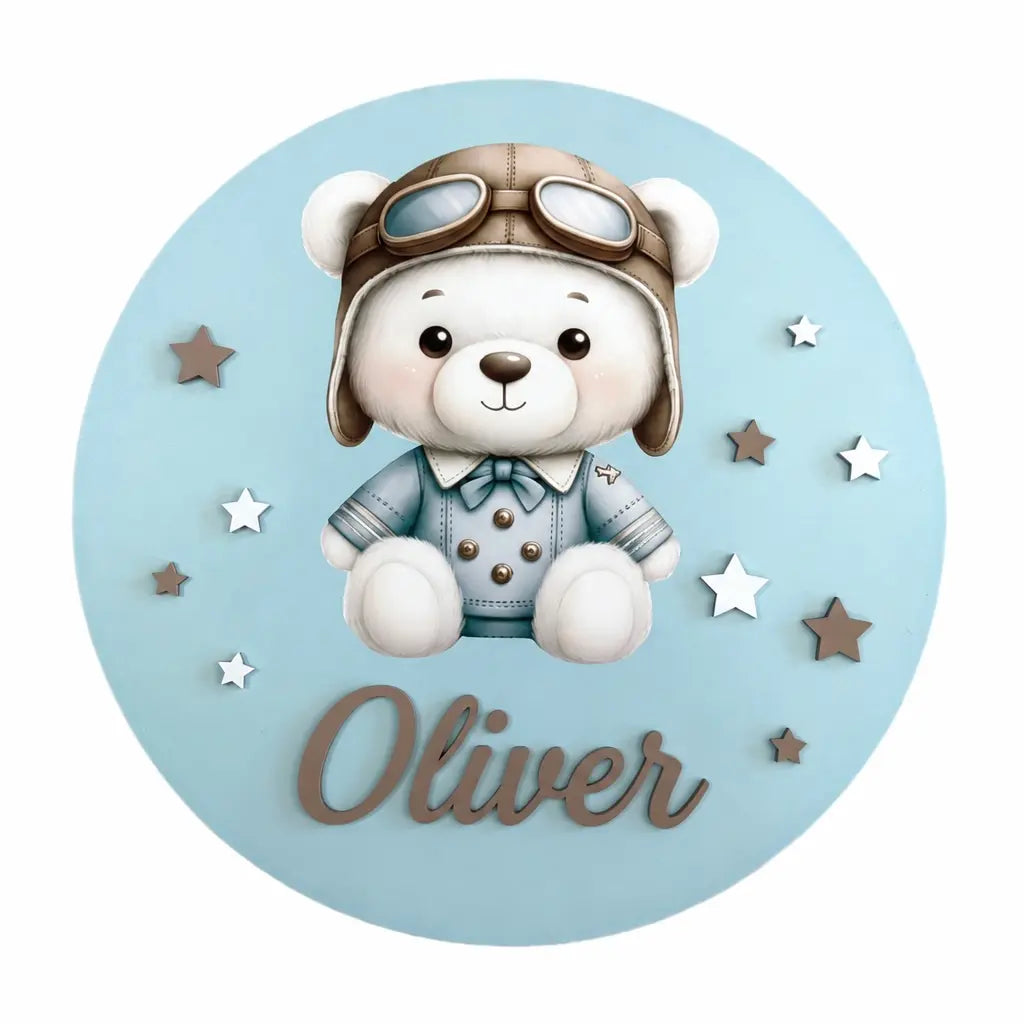 Adorno de madera redondo en azul con un osito aviador, estrellas y el nombre Oliver en relieve.