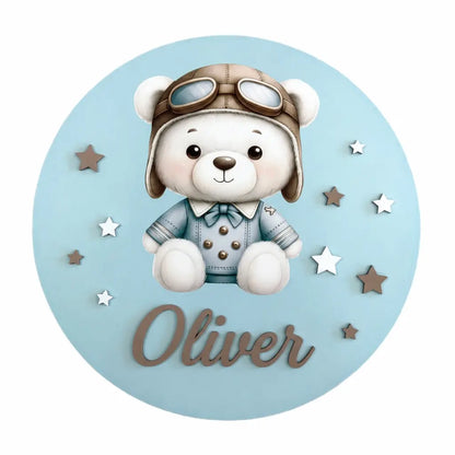 Adorno de madera redondo en azul con un osito aviador, estrellas y el nombre Oliver en relieve.