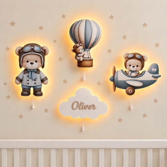 Set de 4 lámparas infantiles de madera Osito volador: osito con gorro de piloto, osito en globo aerostático, osito en avioneta y nube con el nombre Oliver, encendidas en la pared de una habitación de bebé.